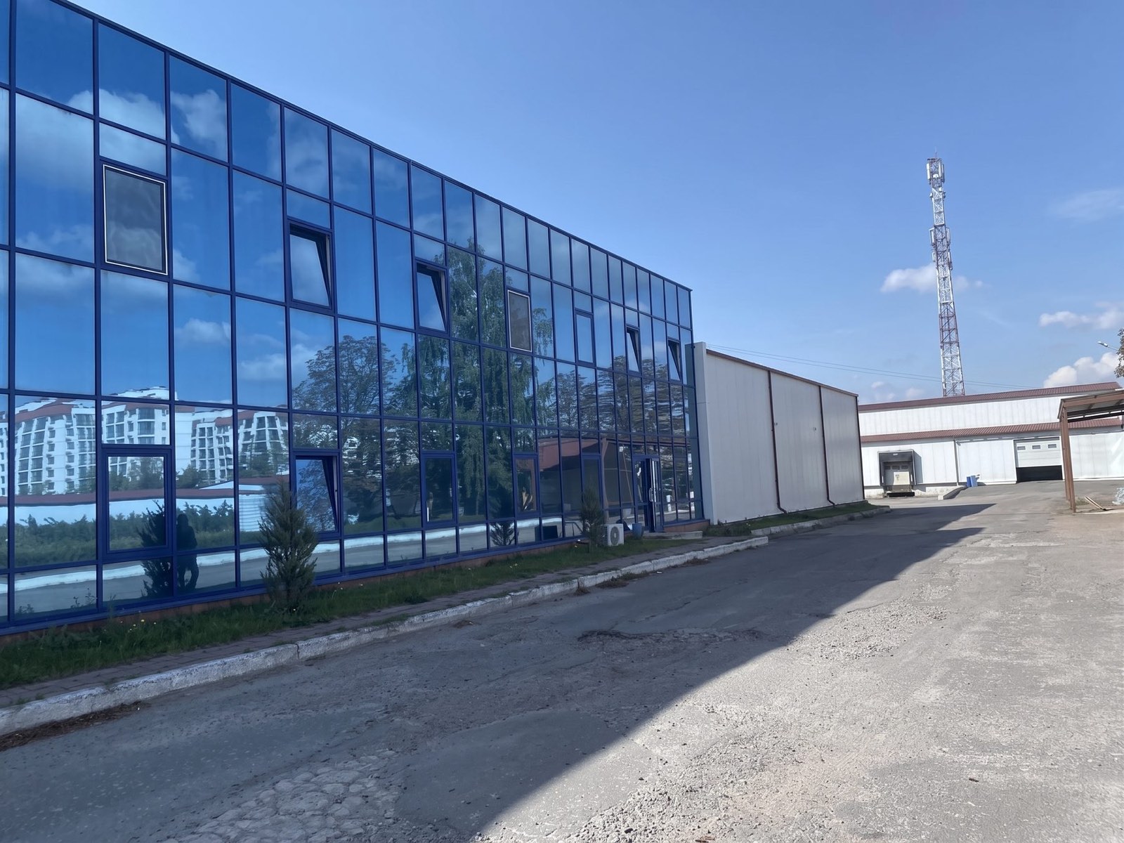 Офісний блок зі суцільним синім склінням на всю довжину фасаду, поруч - глухий торець складського корпусу і внутрішній проїзд між будівлями