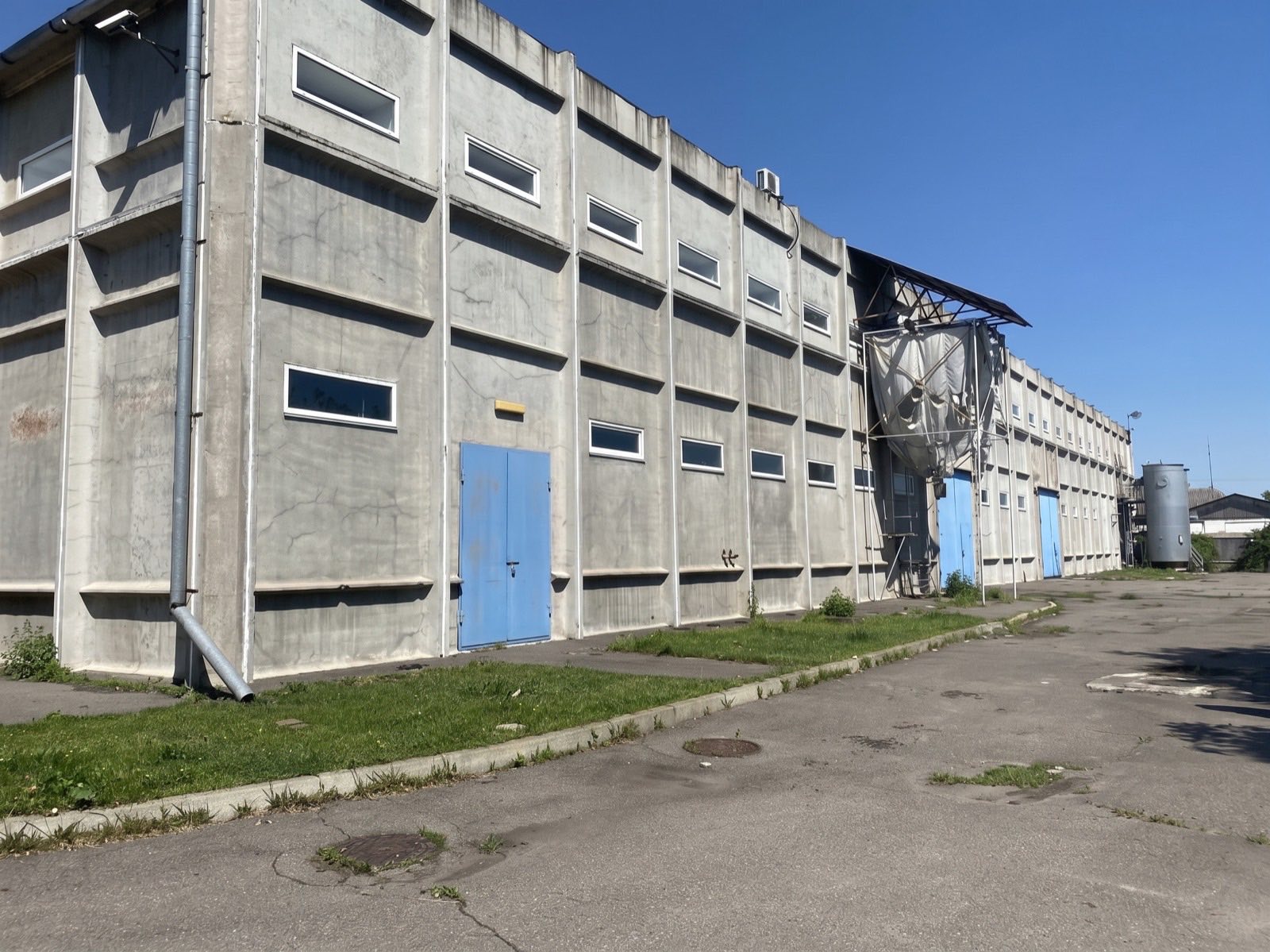 Фасад корпусу з бетонних панелей у два ряди вікон, сині вантажні ворота, праворуч до стіни прибудовані металеві силоси