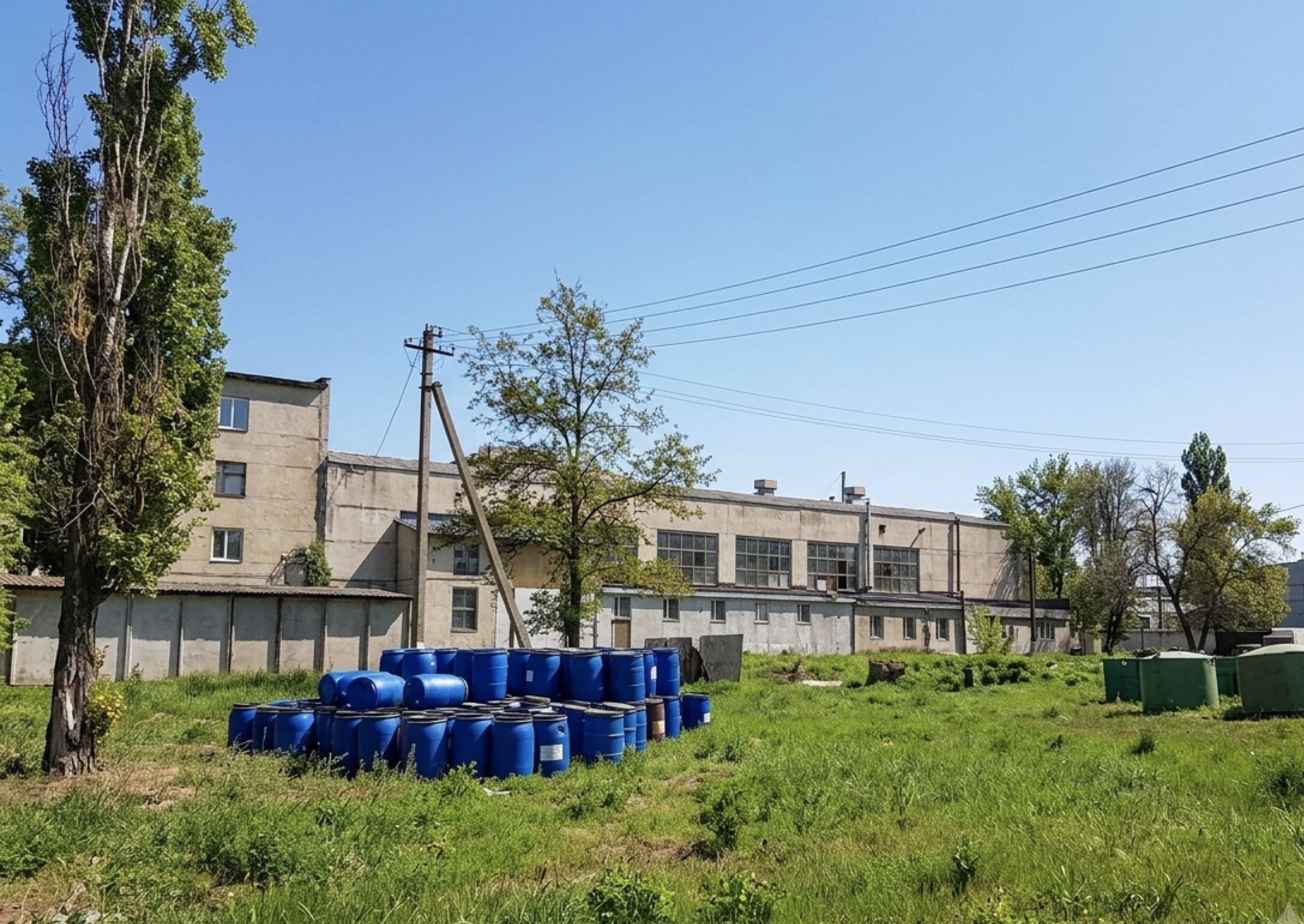 Вільна ділянка території з бочками на майданчику, за нею - корпуси комплексу і бетонний паркан